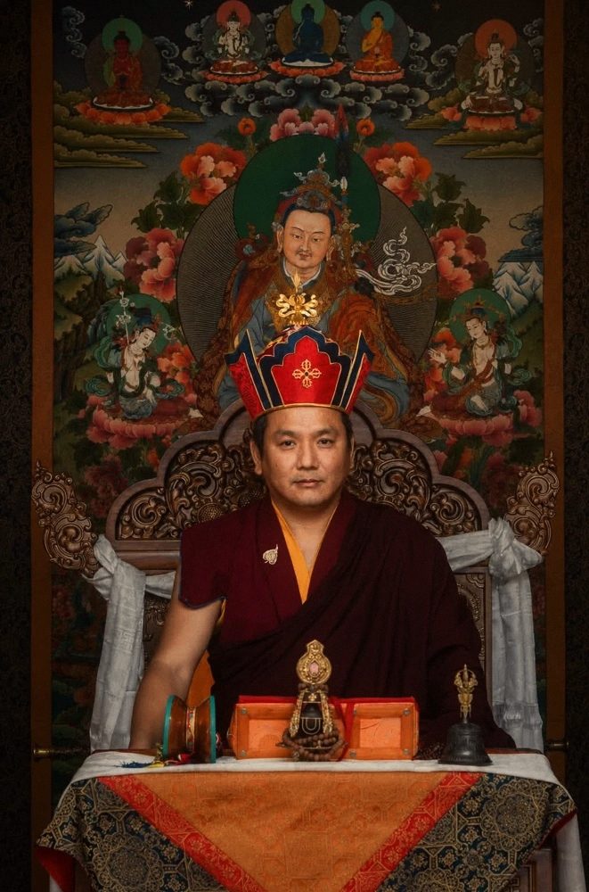 Rinpoche đội mũ đỏ ngồi trước thangka Đức Liên Hoa Sanh